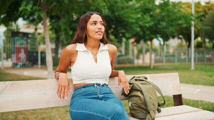 Naklejka premium Lovely tanned woman with long brown hair wearing white top sitting relaxed on park bench looking away. Life style concept. Slow motion.