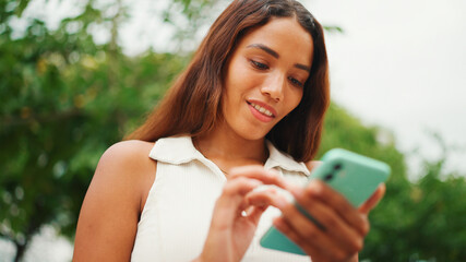 Clouse-up, beautiful girl with long dark hair wearing white top sits on bench and uses mobile phone. Young smiling woman writing in social networks on cellphone.