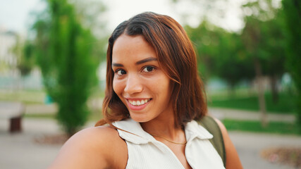 Lovely girl with brown long hair, tanned skin wearing white top is using mobile phone. Smiling young woman taking selfie on cellphone