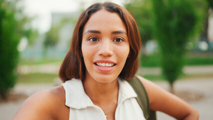 Clouse-up, lovely girl with brown long hair, tanned skin wearing white top is using mobile phone. Smiling young woman taking selfie on cellphone