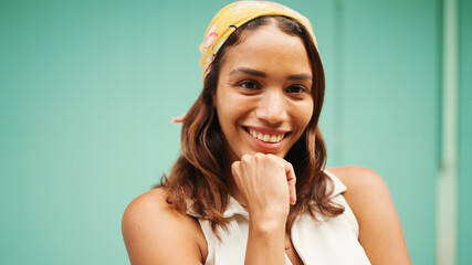 Beautiful girl with tanned skin, brown long hair and bandanna on her head poses for the camera. Smiling young woman putting one finger to the cheek on blue background