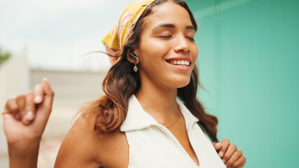 Cute tanned woman with long brown hair wearing white top and yellow bandana listens to music in wireless headphones and dances on blue wall background.