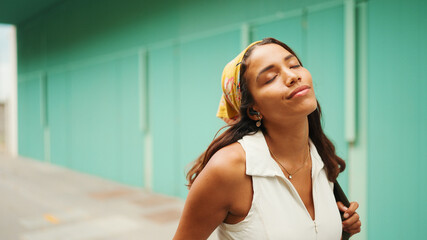 Cute tanned woman with long brown hair wearing white top and yellow bandana listens to music in wireless headphones and dances on blue wall background.