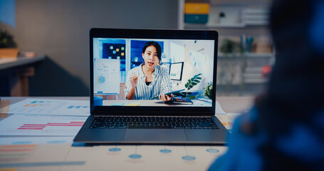 Asia woman using laptop talk to colleagues about work in video call while working from home at living room at night. Self-isolation, social distancing, quarantine for coronavirus prevention.