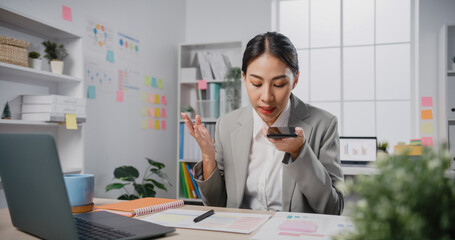 Young Asia cheerful professional woman  sitting on desk with laptop record audio voice message on smartphone confirm detail at office. Lady business suit, Technology startup business concept.