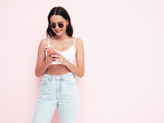 Young beautiful smiling female in trendy summer clothes. Sexy carefree woman posing near pink wall in studio. Positive brunette model looking at cellphone screen. Holding smartphone and using apps