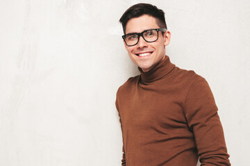 Fototapeta premium Portrait of handsome smiling model. Sexy stylish man dressed in turtleneck sweater and jeans. Fashion hipster male posing near grey wall in studio. Isolated. In spectacles or eyeglasses