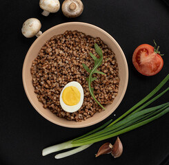buckwheat porridge with egg in a clay bowl with mushrooms, green onion and tomato on a black background