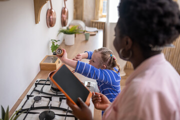 A woman holding a tablet while a girl stirrinf something in a bowl on the gas stove