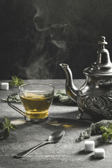 Traditional Moroccan teapot with a steaming cup of tea in a smoky black atmosphere. Vertical format.