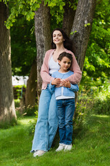 Fototapeta premium full length of positive woman hugging daughter near trees in park