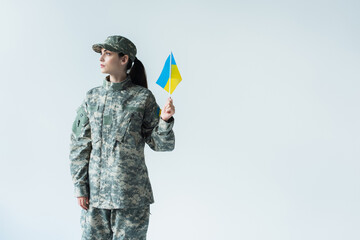 Soldier in uniform and cap holding ukrainian flag and looking away isolated on grey