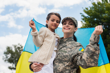 happy military woman with daughter and ukrainian flag looking at camera outdoors