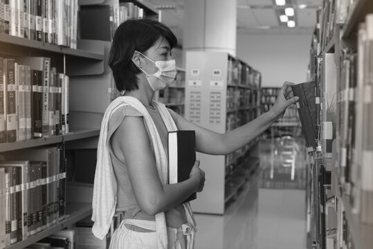 Woman with face mask searching data and select book for research on shelf