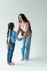 full length of cheerful woman holding hands of daughter with pigtails on grey background