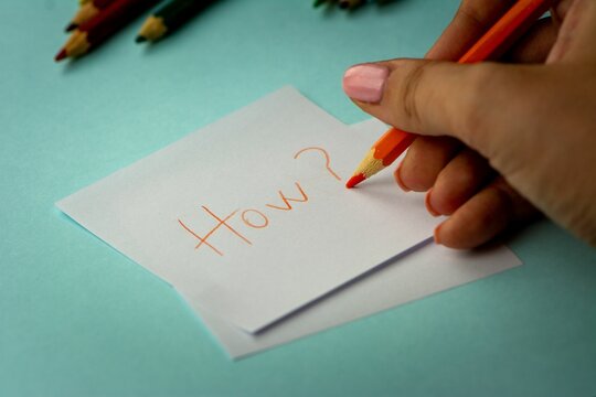 Women's hand writing how on a piece of paper with some other pencils in the background
