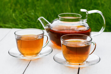 Tea in glass teapot and two cups on a wooden white background background against the background of green grass. Teapot with green or black tea. Tea time