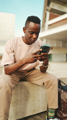 Young african student sits outside of university, uses cellphone, taps on smartphone screen