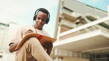 Young african student sitting outside of university and taking notes while study online with...