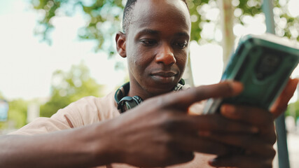 Smiling young african student sits on bench outside of university in headphones, uses phone