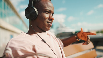 Smiling young african guy relaxes sitting on bench in the port, listens to music in headphones,...
