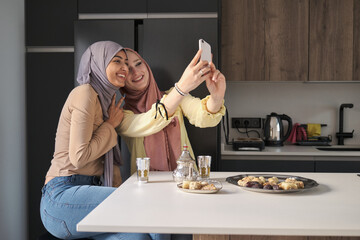 Two muslim women taking a selfie while having tea and arabic sweets at the kitchen table.