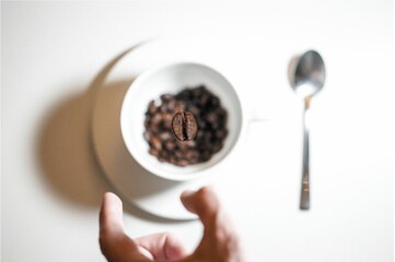 Top view of a man's hand tossing a large coffee bean into a white coffee cup with a spoon beside it