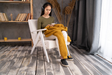 woman wearing headphones knitting at home, sitting on cozy armchair in living room, happy positive...