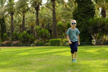 The boy jumps for joy on a green field. A child in sunglasses on a background of palm trees. Concept: holidays, travel, exotic destinations, tropical countries.