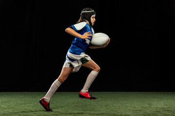 Dynamic portrait of school age boy, junior male rugby player practicing rugby football isolated on dark background with grass floooring.