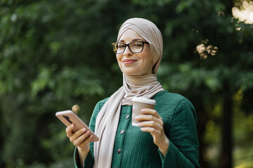 Attractive smiling muslim woman in hijab texting on smart phone standing with hot drink on background of green city park. Portrait of young arabian businesswoman using mobile phone outdoor. - Powered by Adobe