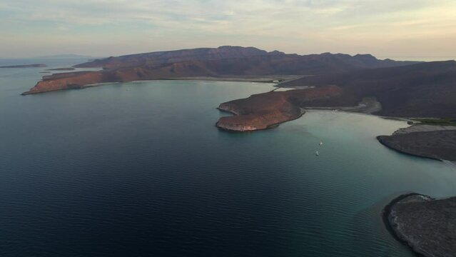 Aerial view of the wavy sea and the rocky mountains gleaming under the cloudy blue sky in 4K