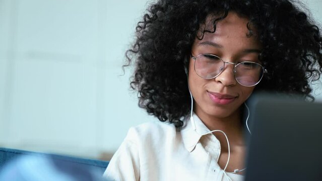 Laughing curly haired woman working on laptop in headphones while sitting on the sofa at home 