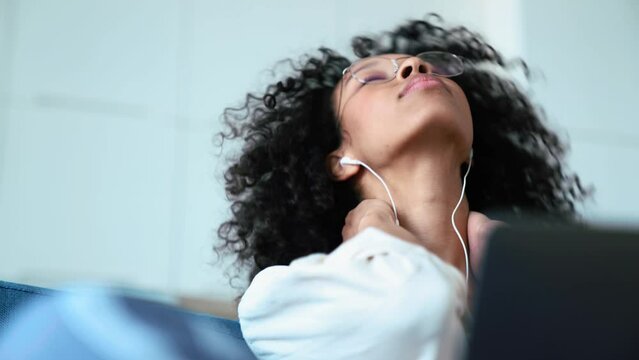 Tired curly haired woman working on laptop in headphones and stretching while sitting on the sofa at home 