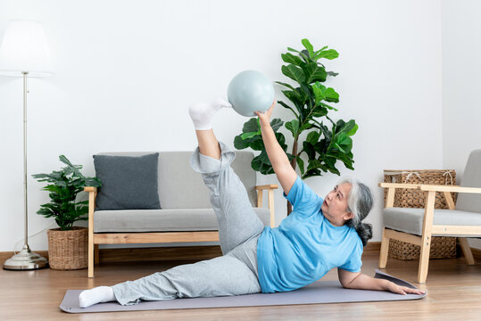 Asian Elderly Woman Doing Exercise At Home By Stretching The Leg Muscles And Using A Ball As An Exercise Aid, To People Retirement Age And Health Care Concept.