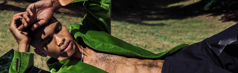 fashionable african american man in green velvet blazer lying on lawn with hands near head, banner