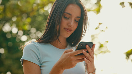 Young beautiful smiling woman with long brown hair wearing white crop top sitting in park having picnic on summer day outdoors, talking with friends, using cellphone