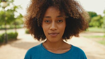 Close-up of young smiling woman with curly hair wearing blue t-shirt posing for the camera in the park. The girl opens her eyes and smiles at the camera