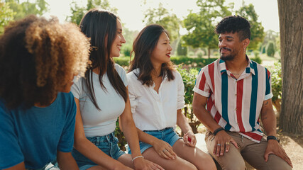 Happy multiethnic young people talking while sitting on park bench on summer day outdoors. Group of friends talking and laughing merrily in city park