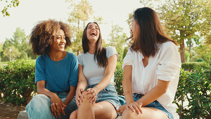 Happy multiethnic young women talking while sitting on park bench on summer day outdoors. Group of girls talking and laughing merrily in city park