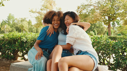 Naklejka premium Happy multiethnic young women talking while sitting on park bench on summer day outdoors. Group of girls talking and laughing merrily in city park