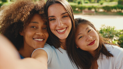 Happy, lovely multiethnic young women posing on cellphone camera on summer day outdoors. group of friends hugging each other smiling at the camera while standing on path in the park