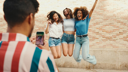 Happy, lovely multiethnic young people posing for the camera taking photo on summer day outdoors. Young man photographing smiling girls on cellphone camera