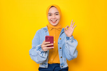 Happy young Asian woman in jeans jacket holding mobile phone and showing okay sign isolated over yellow background