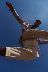 bottom view of african american man in white pants jumping under blue sky