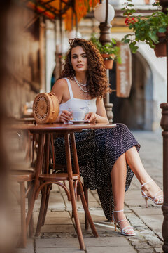 Fototapeta Elegant curly brunette woman drinking coffee in street cafe in European city. Summer vacation, travel, coffee break, lifestyle conception. Full-length outdoor portrait 