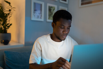 Focused black young man works on computer remotely from home sitting in living room at night. African American programmer writes code for website looking at screen of modern electronic device closeup