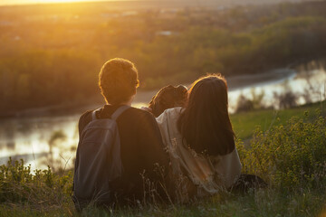 a young guy and a girl with their big dog sit on a high bank, a river flows below and enjoy the view of sunset nature on vacation during a trip