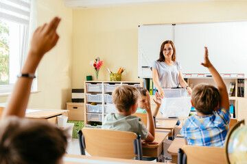 Save the earth. Charming female teacher with small school children in classroom learning about waste separation. Children raised their hands to answer, rear view.