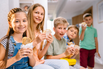 Different age pupils in school corridor during lunch break. Hungree children eating snack inside.